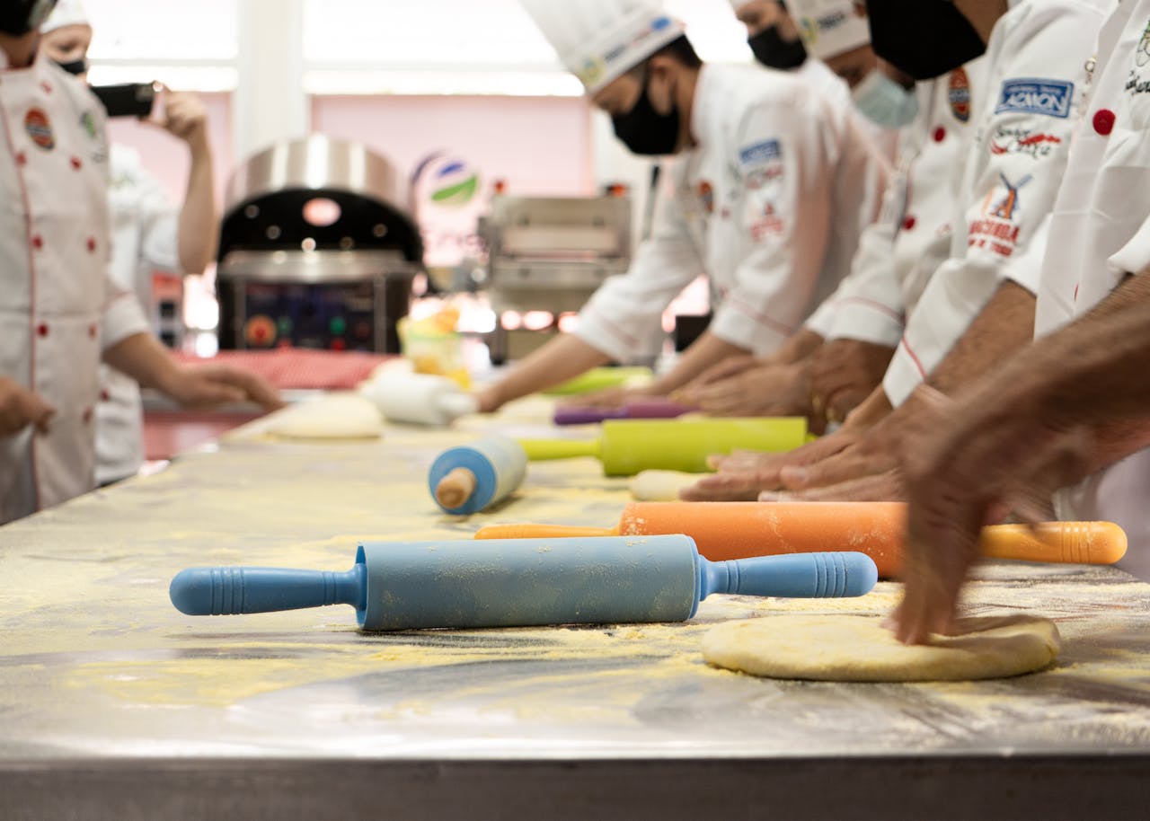 service-02 Chefs wearing uniforms and masks prepare dough with colorful rolling pins in a professional kitchen.