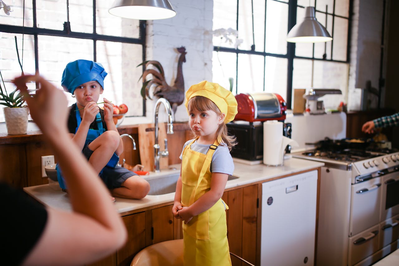 service-03 Two kids in colorful chef hats enjoy a playful cooking session in a cozy kitchen setting.