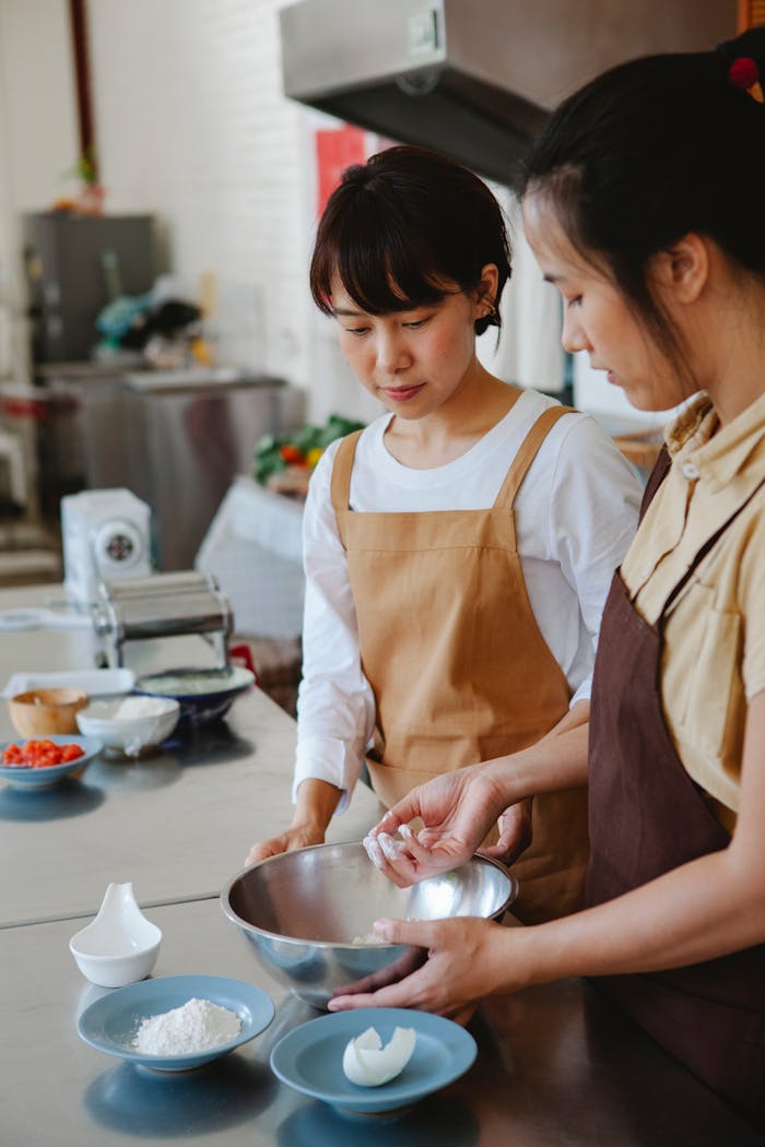 services-img Two Asian women baking together in a modern kitchen, preparing dough with fresh ingredients.