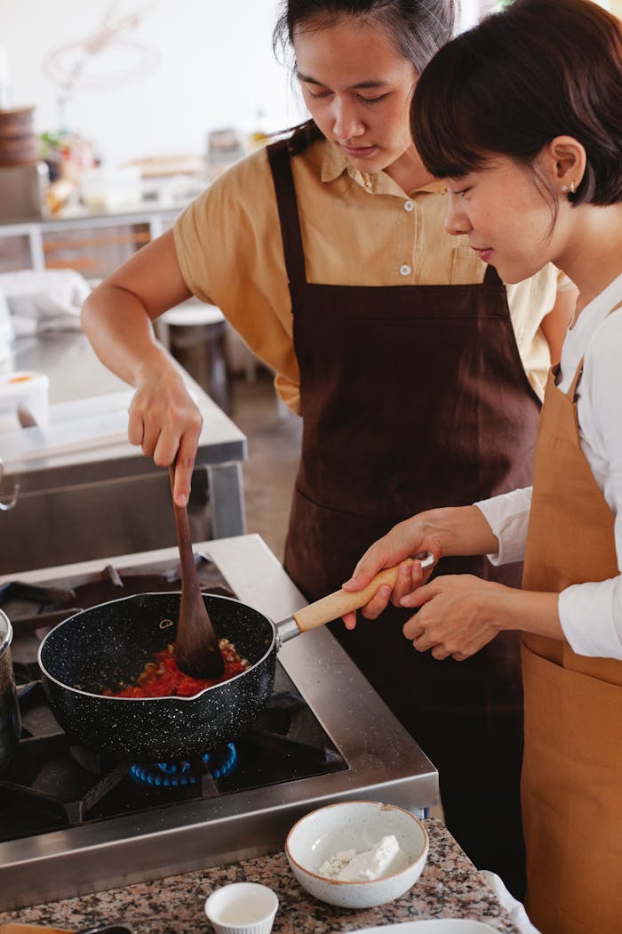 my-steps Two women wearing aprons cook together in a modern kitchen, focusing on a pan dish.