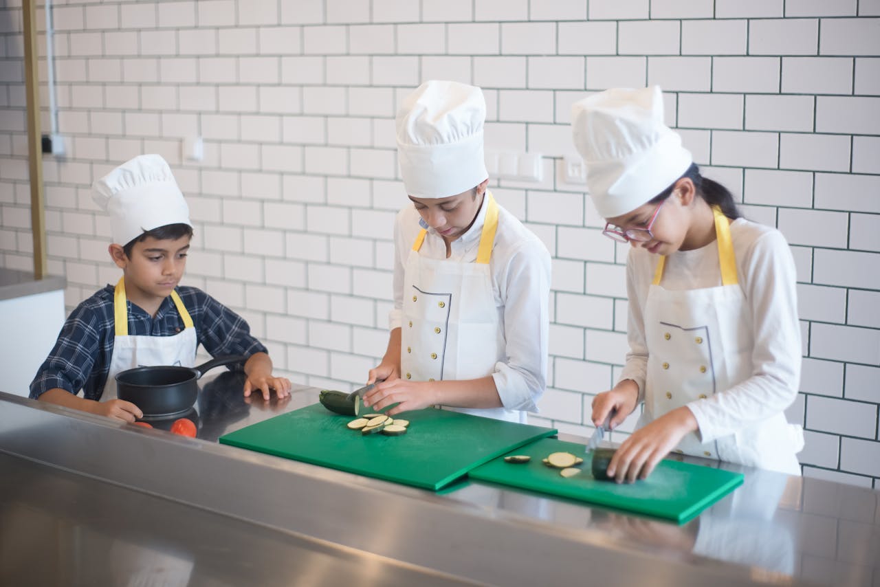 about-me-img Kids wearing chef hats and aprons cooking in a professional kitchen setting.