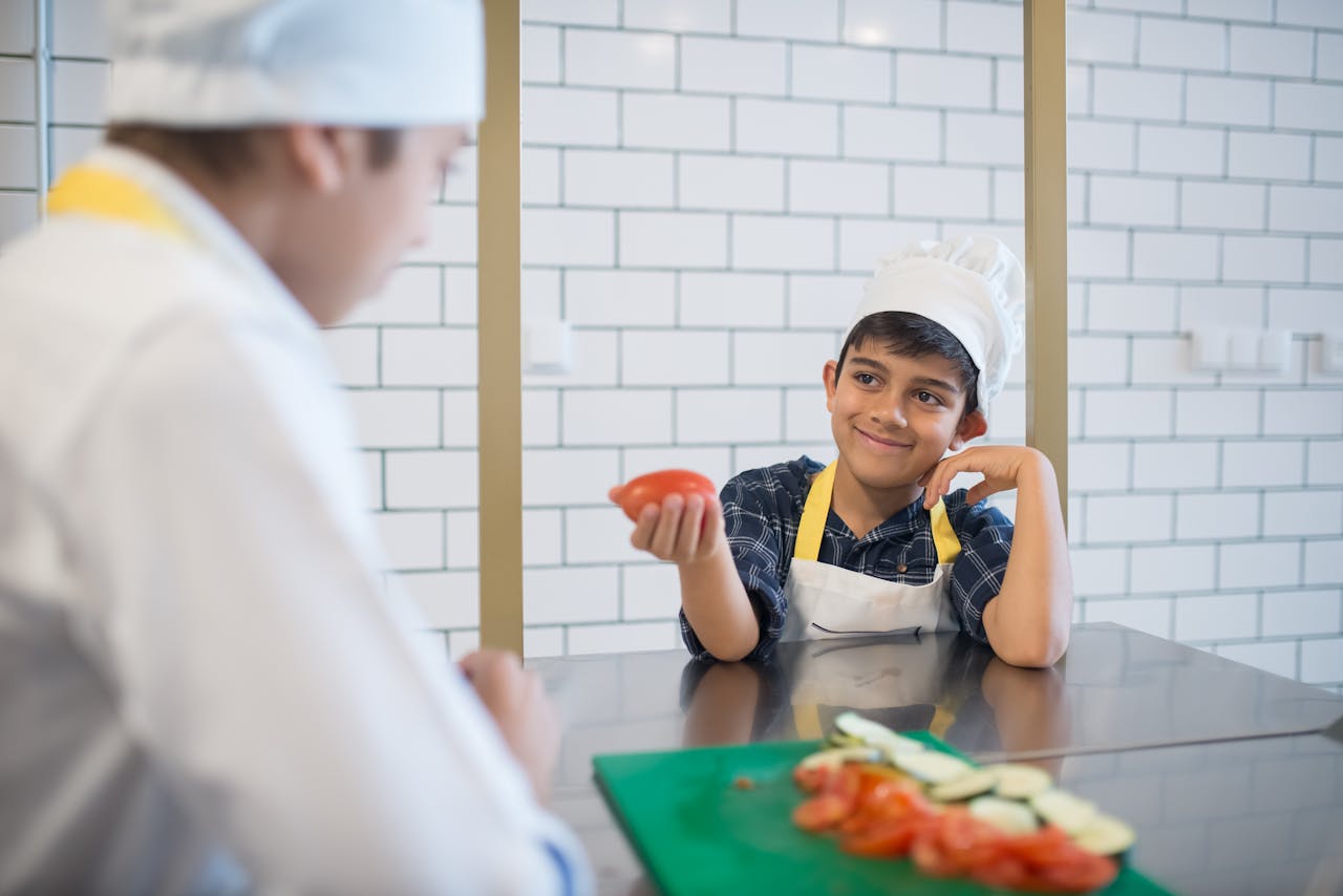 why-choose-me Smiling young boy wearing a chef's hat in a kitchen, proudly holding a tomato.